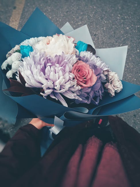 A vibrant bouquet of fresh flowers held by a person, featuring a mix of pastel pink roses, large white and purple chrysanthemums, smaller blue accents, and greenery, arranged in a stylish dark blue wr