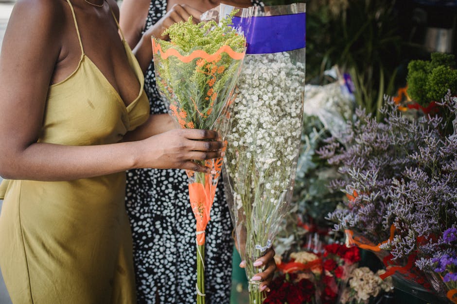 A woman dressed in a yellow satin slip dress holds a bouquet of green and purple flowers with small clusters of blooms, wrapped in clear cellophane, in a flower shop. Her hand gently cradles the bouqu
