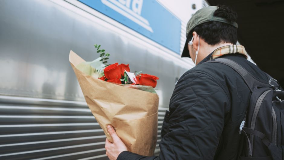 A person wearing a grey cap, black jacket, and a backpack, holding a bouquet of fresh red roses wrapped in brown craft paper, is captured mid-action near a silver train carriage. The bouquet features 