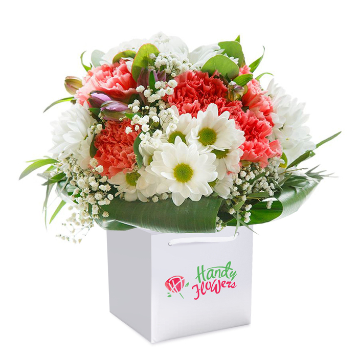 Graceful and inviting, this vibrant floral arrangement-a favourite among Motspur Park residents-is presented upright in a strikingly crisp, white square gift bag designed for both style and ease of delivery. Every element is composed with the practiced hand of a local florist, its palette evoking the soft morning light of parks near Motspur Park station. At the bouquet's heart, coral-toned carnations unfold in textured, ruffled layers, while blush and scarlet varieties mingle for a sense of lively abundance. White daisies pop throughout, their golden-green centers gleaming with a joyful brightness reminiscent of springtime wildflowers on Adela Avenue. Delicate, cloud-like sprays of baby's breath thread the arrangement, imparting subtle lightness and air. Close by, nuanced hints of unopened blossoms tinged with gentle purple add a layer of mystery and freshness, suggestive of new beginnings. The ensemble is surrounded by strong, glossy leaves that arch in lush, protective swaths, framing the flowers and forming a beautifully rounded silhouette. The bag's discrete white ribbon handles and elegant simplicity compliment the presentation, while a seamless white background focuses attention solely on the bouquet's details. Every bloom suggests care, expertise, and the promise of freshness-an ideal choice for birthdays, anniversaries, or heartfelt gifts shared locally within Motspur Park, crafted by hands that know the community intimately.