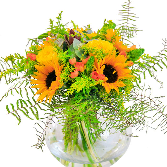 The first impression this bouquet conjures is radiant joy-an abundant mix of yellow and orange blooms set against fresh greenery, sitting proudly in a round, gleaming glass vase. Large, cheerful sunflowers with deep brown centers anchor the arrangement, echoing the sun-drenched fields surrounding Motspur Park during the height of summer. Woven between are bright yellow garden roses, petals unfolding in tight, luxurious spirals, their soft texture inviting both admiration and touch. Tiny, glossy red Hypericum berries peek out from the fronds, providing lively contrast and a touch of playful whimsy. Feathery green ferns and slender sprays of light green solidago give the bouquet movement, curling around the arrangement's edges as if dancing in a gentle breeze drifting over Prince George's Playing Fields. Subtle, blue-tinged eucalyptus leaves add both texture and a refreshing note to the ensemble. The clear glass vase-its elegant, rounded shape reminiscent of a classic fishbowl-houses a lush tangle of stems, magnifying the bouquet's natural architecture and sustaining its vibrancy in pure water. Crafted with the care of a skilled Motspur Park florist, this bouquet's sunlit exuberance is perfect for birthdays, anniversaries, or brightening the day of someone dear, echoing the spirit and togetherness found in the local community.